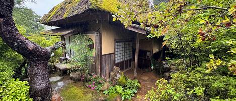 A traditional Japanese house with a thatched roof, surrounded by lush greenery and a small stream.