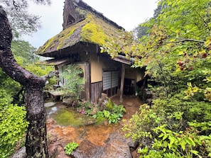Una casa japonesa tradicional con techo de paja, rodeada de abundante vegetación y un arroyo pequeño.
