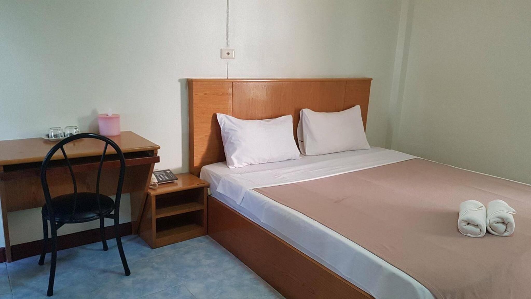 Une chambre avec une tête de lit en bois, un lit simple avec des oreillers blancs, un bureau avec une chaise, une petite table avec un récipient rose, et une paire de serviettes enroulées posées sur le lit.