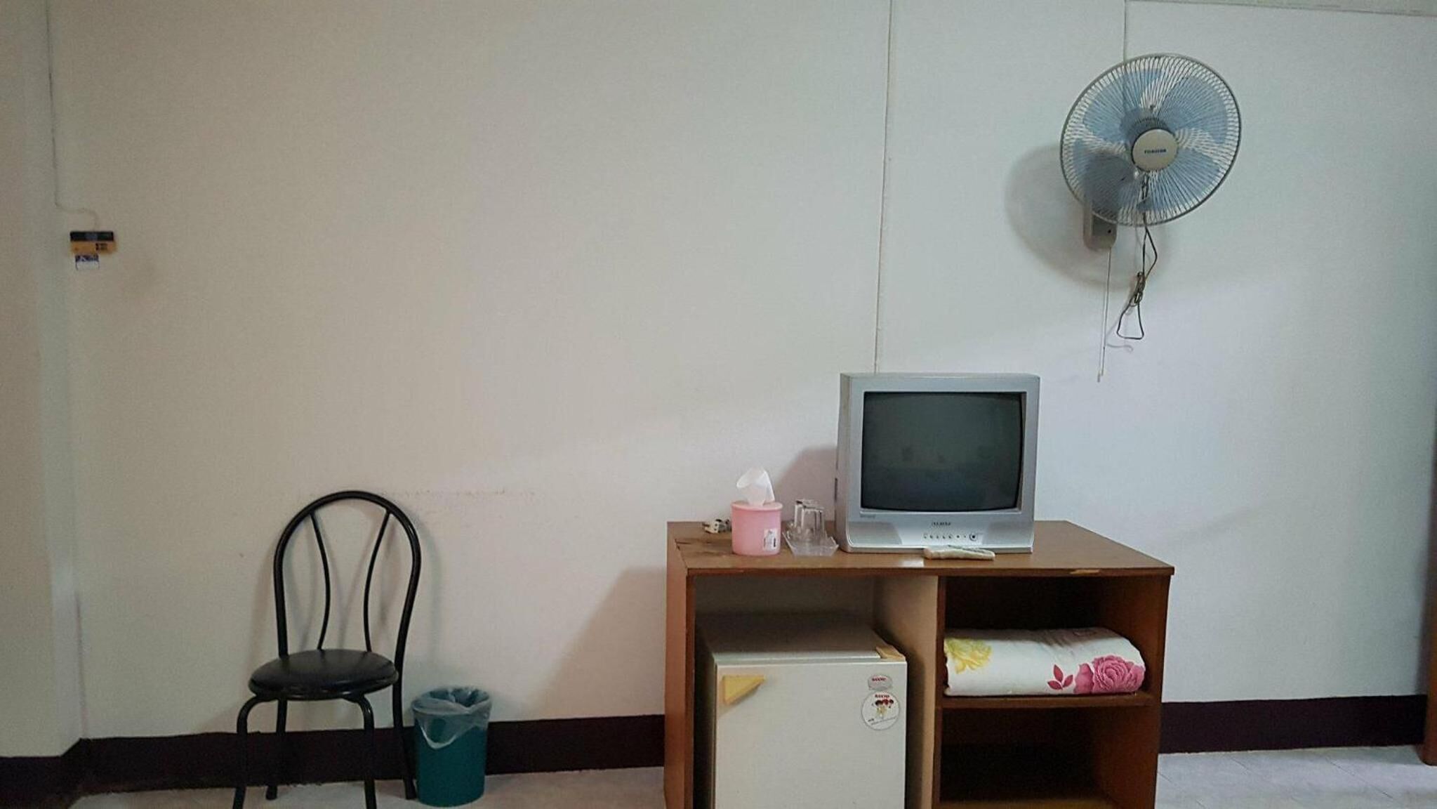 Una habitación con un escritorio de madera, un televisor, una silla, un cesto de basura y un ventilador de pared.