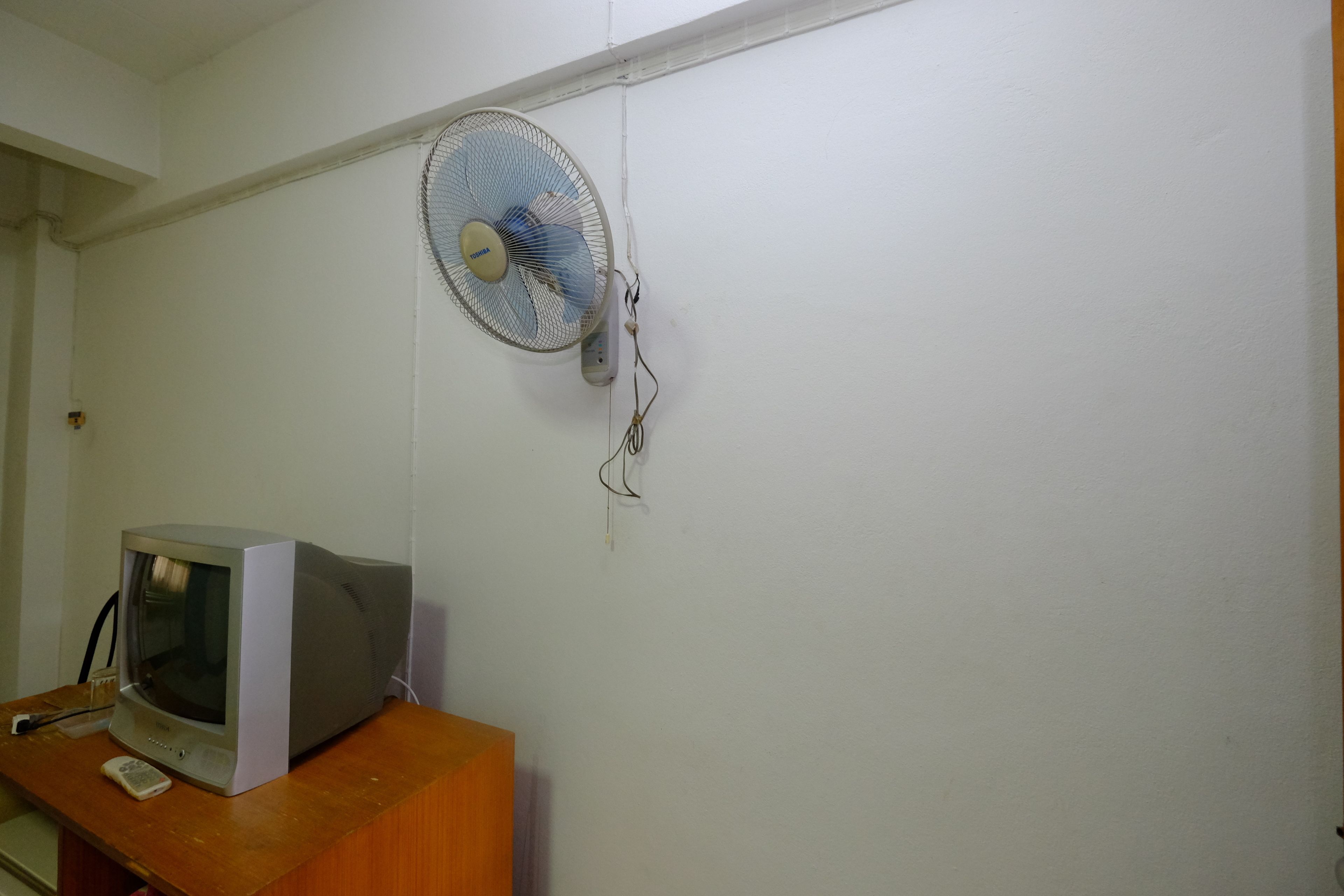 Una habitación con un escritorio de madera, televisor y un ventilador en la pared.