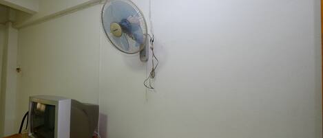 Una habitación con un escritorio de madera, televisor y un ventilador en la pared.