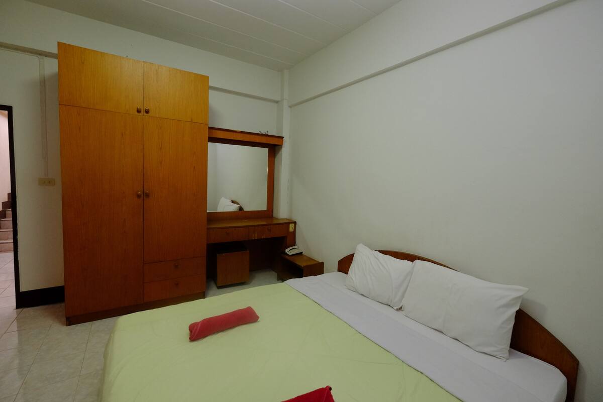 A hotel room with a bed, a wooden wardrobe, a desk with a mirror, and a small bedside table.