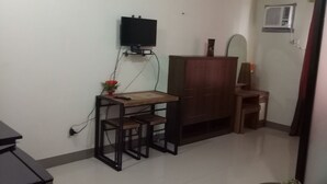 Een kleine, eenvoudig ingerichte kamer met een houten tafel en stoelen, een aan de muur bevestigde televisie en een klein kastje.