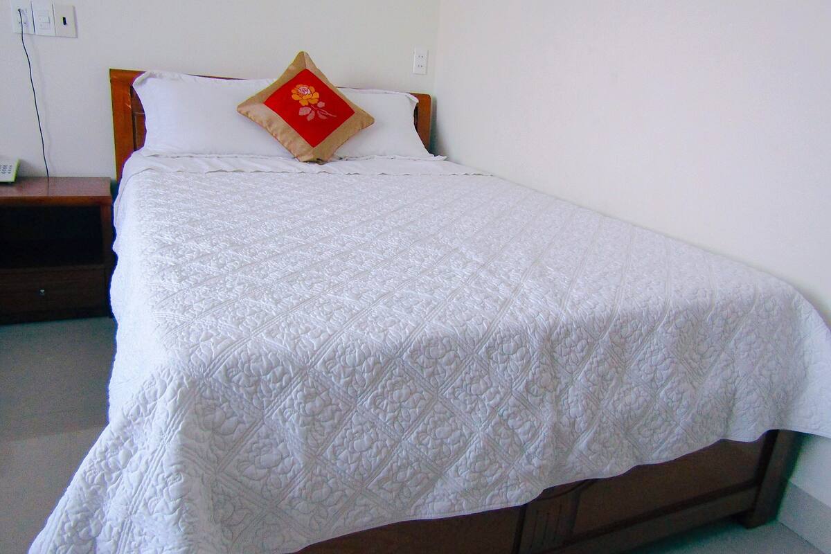Una cama bien hecha con colcha blanca, cabecera de madera y almohada roja con diseño floral.
