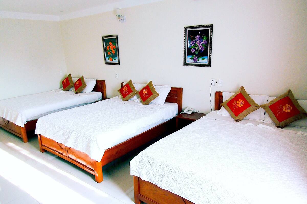 Una habitación de hotel con tres camas individuales, cada una con cabecero de madera y ropa de cama blanca. Almohadas decorativas con diseños florales en cada cama.