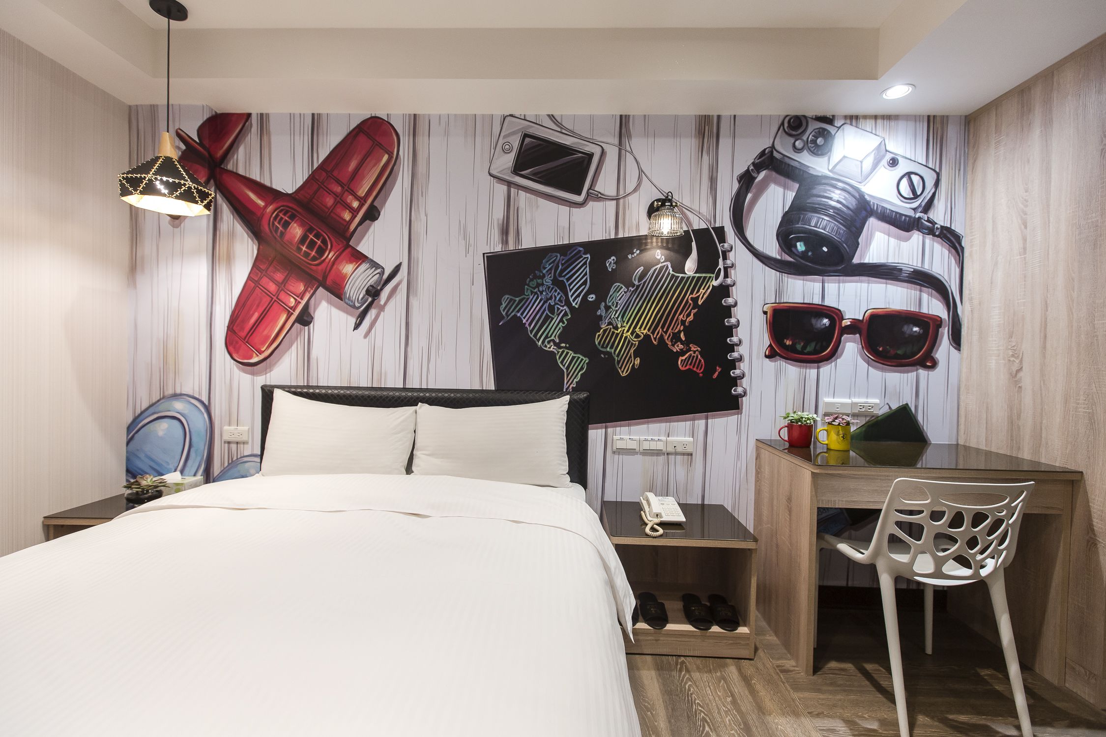 A hotel room with a bed, a desk, and a chair. There are wall decorations including a red airplane, a camera, and sunglasses.