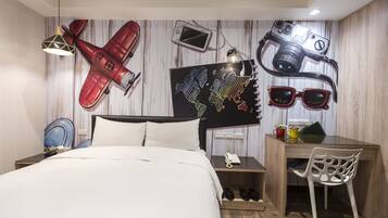 A hotel room with a bed, a desk, and a chair. There are wall decorations including a red airplane, a camera, and sunglasses.