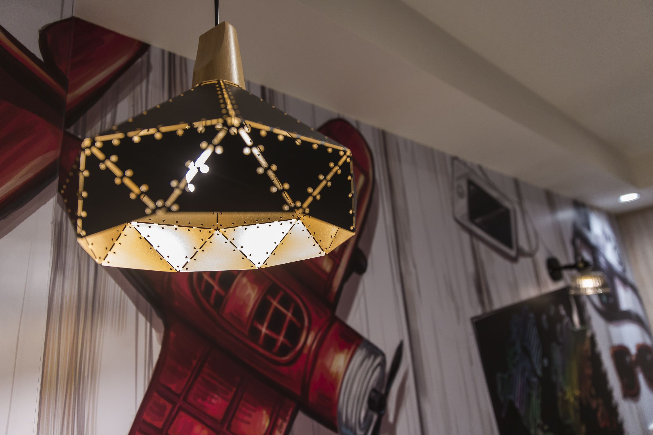 Una lámpara colgante de diseño geométrico con detalles negros y dorados, ubicada en una habitación con un mural de un teléfono rojo.