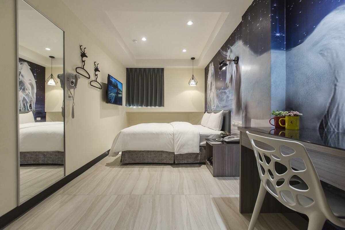 A modern bedroom with a bed, a desk, a chair, a mirror, and a wall with a painting.