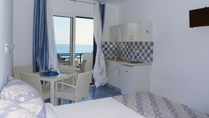 A hotel room with a bed, a small table, a chair, a desk, a window with curtains, a checkered backsplash, and a view of the sea. - Bouganville Studios (Rhodes)