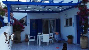 Un área de estar al aire libre cubierta con sillas blancas, una mesa, una pérgola azul y plantas en macetas.