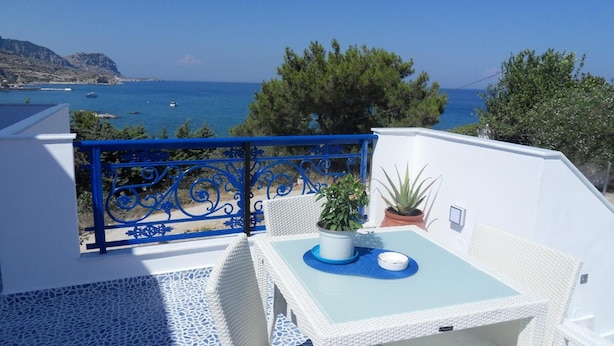 A balcony with a white wicker table and chairs, a potted plant, and a view of the sea and mountains. - Bouganville Studios (Rhodes)
