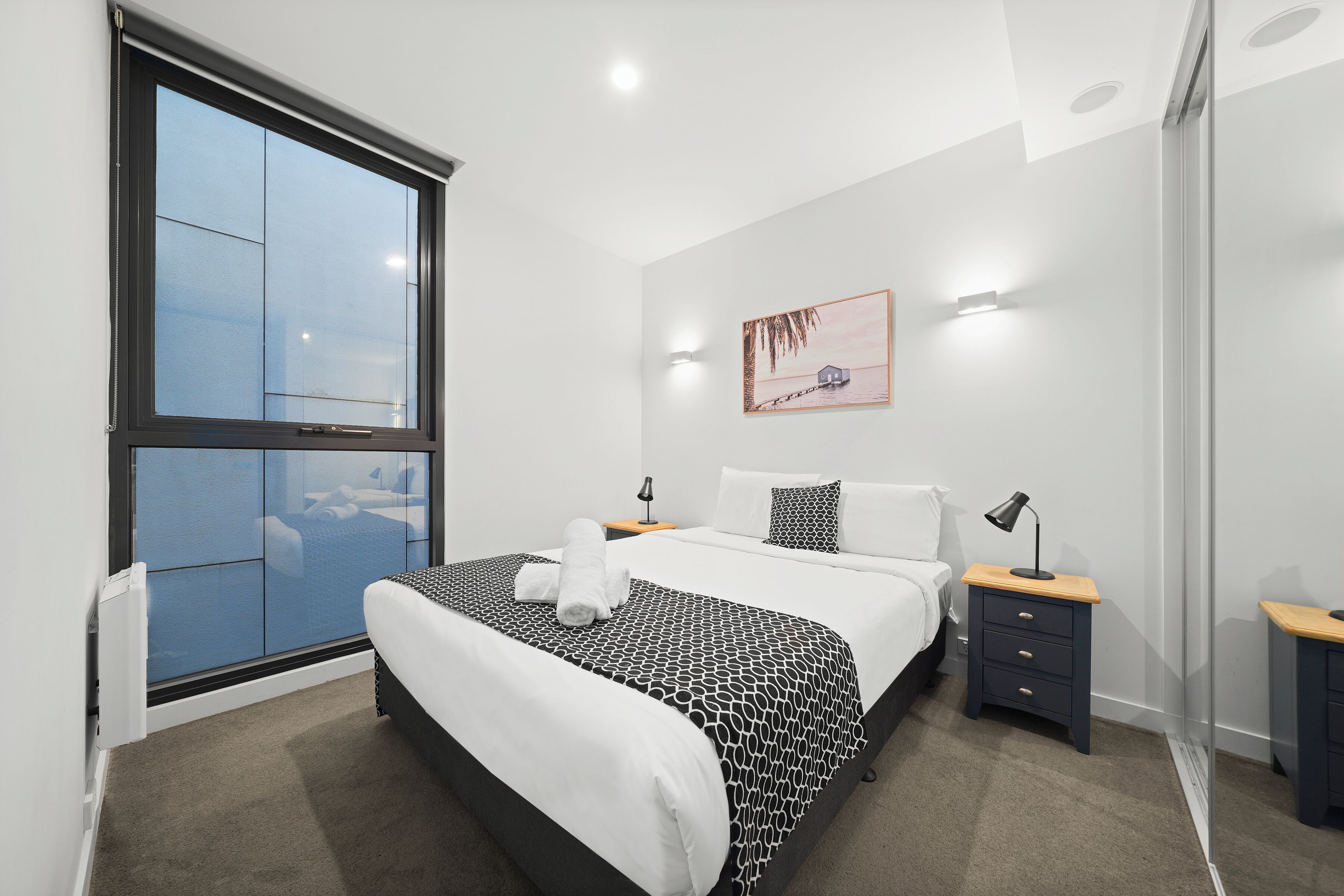 A modern bedroom with a large window, a bed with a patterned bedspread, a bedside table with a lamp, and a wall-mounted artwork.