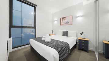 A modern bedroom with a large window, a bed with a patterned bedspread, a bedside table with a lamp, and a wall-mounted artwork.