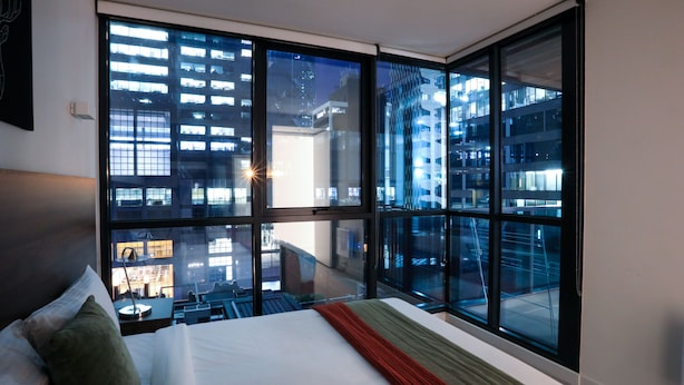 Une chambre d’hôtel dotée d’une grande fenêtre donnant sur la ville, d’un lit parfaitement fait et d’un design moderne.