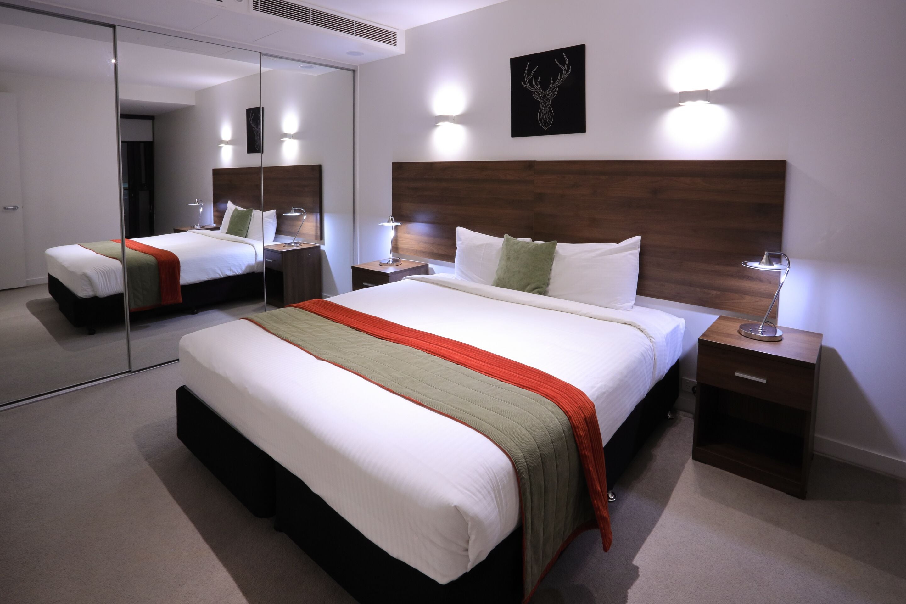 A modern hotel room with a large bed, bedside tables, a mirror, and a wall-mounted light fixture.