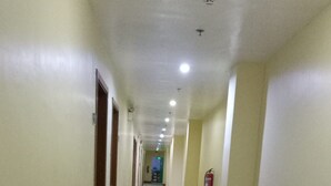 A long, well-lit hallway with doors on both sides, a fire extinguisher on the right wall, and a tiled floor.