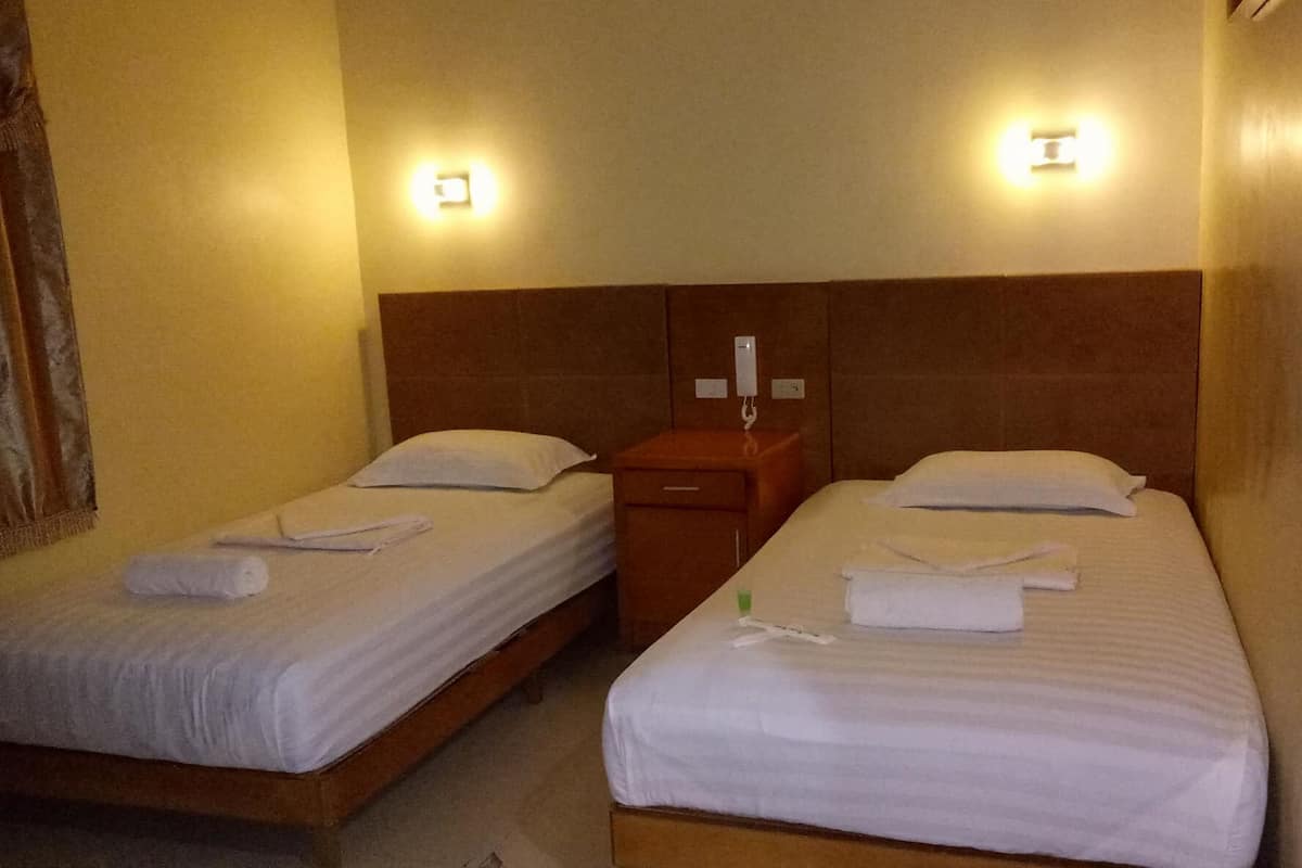 A hotel room with two single beds, each with a pillow and a rolled towel. There is a wooden nightstand between the beds with a phone and a small drawer.