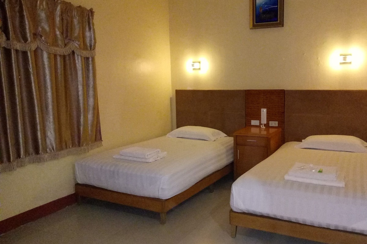 A hotel room with two single beds, each with a pillow and a folded towel. There is a nightstand between the beds, and a wall-mounted light fixture above each bed.