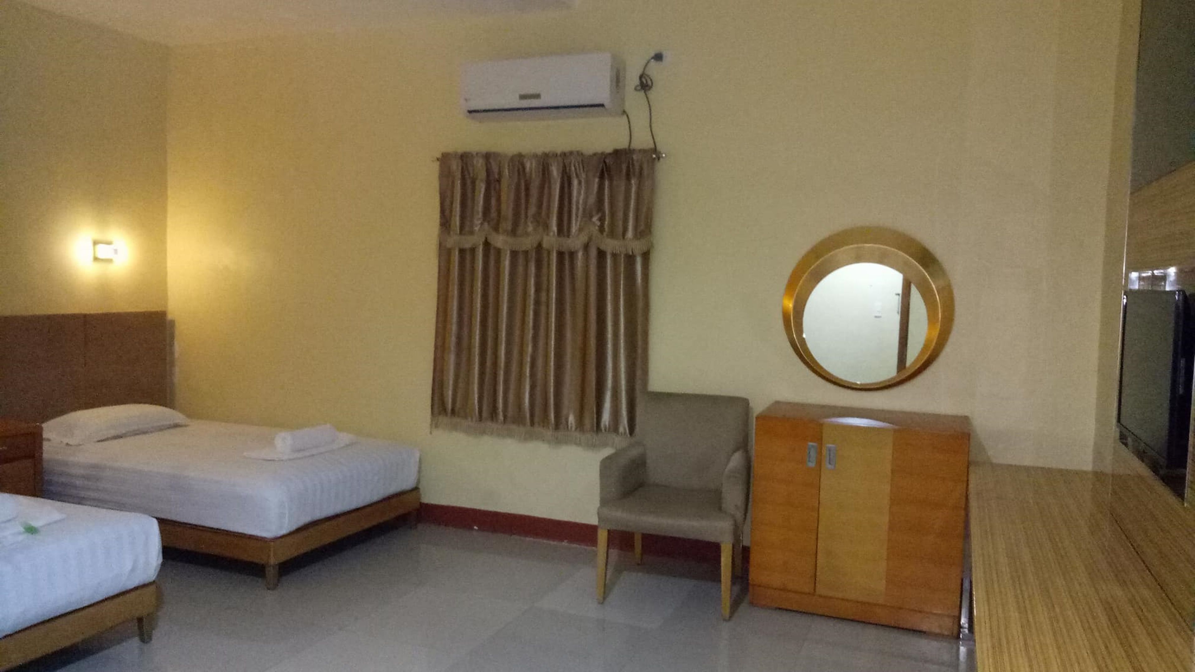 A hotel room with two single beds, a wooden wardrobe, a chair, a round mirror, and an air conditioning unit on the wall.
