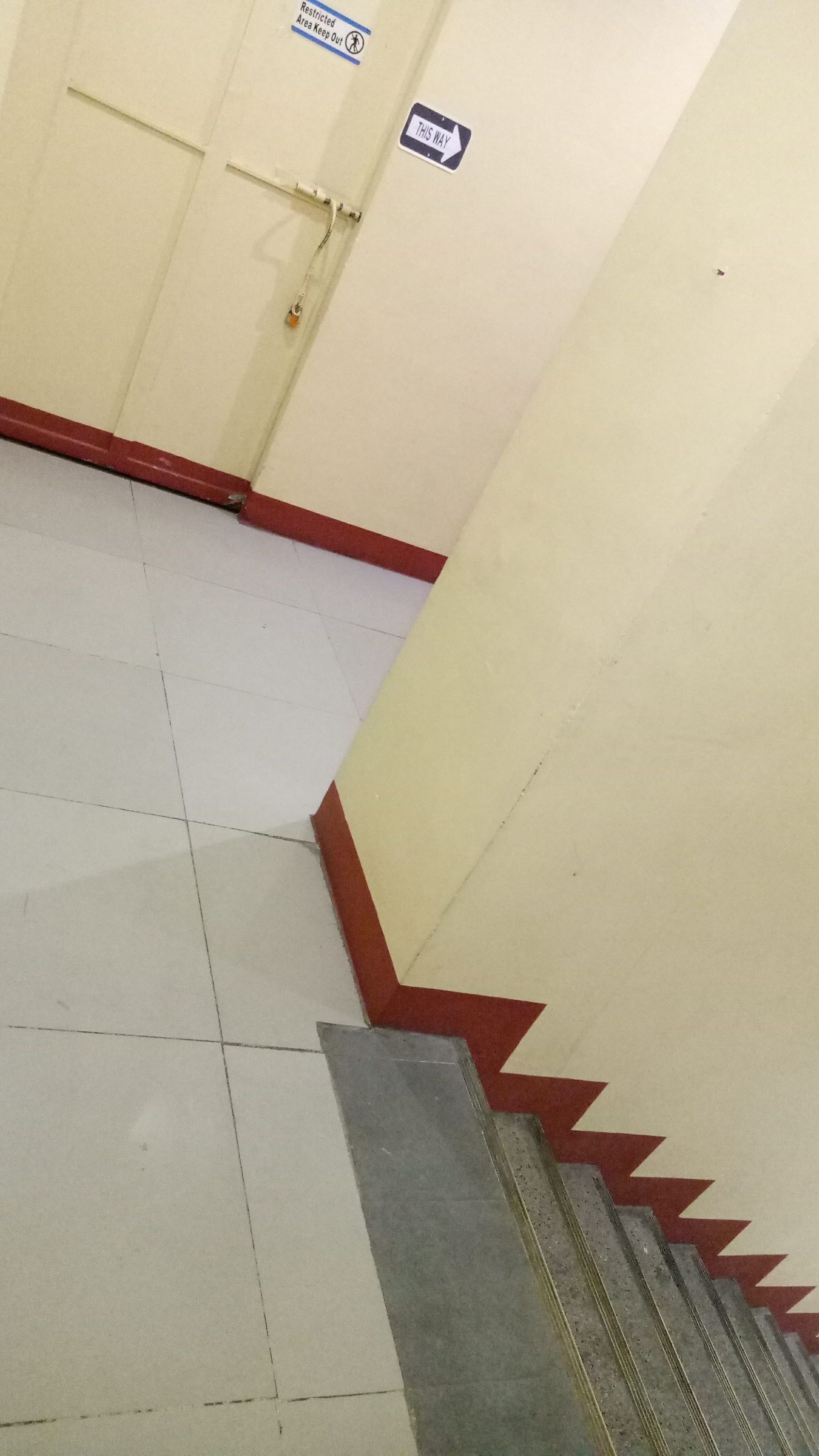 A corridor with a tiled floor, a door with a 'Restricted Area Keep Out' sign, and a 'This Way' sign.