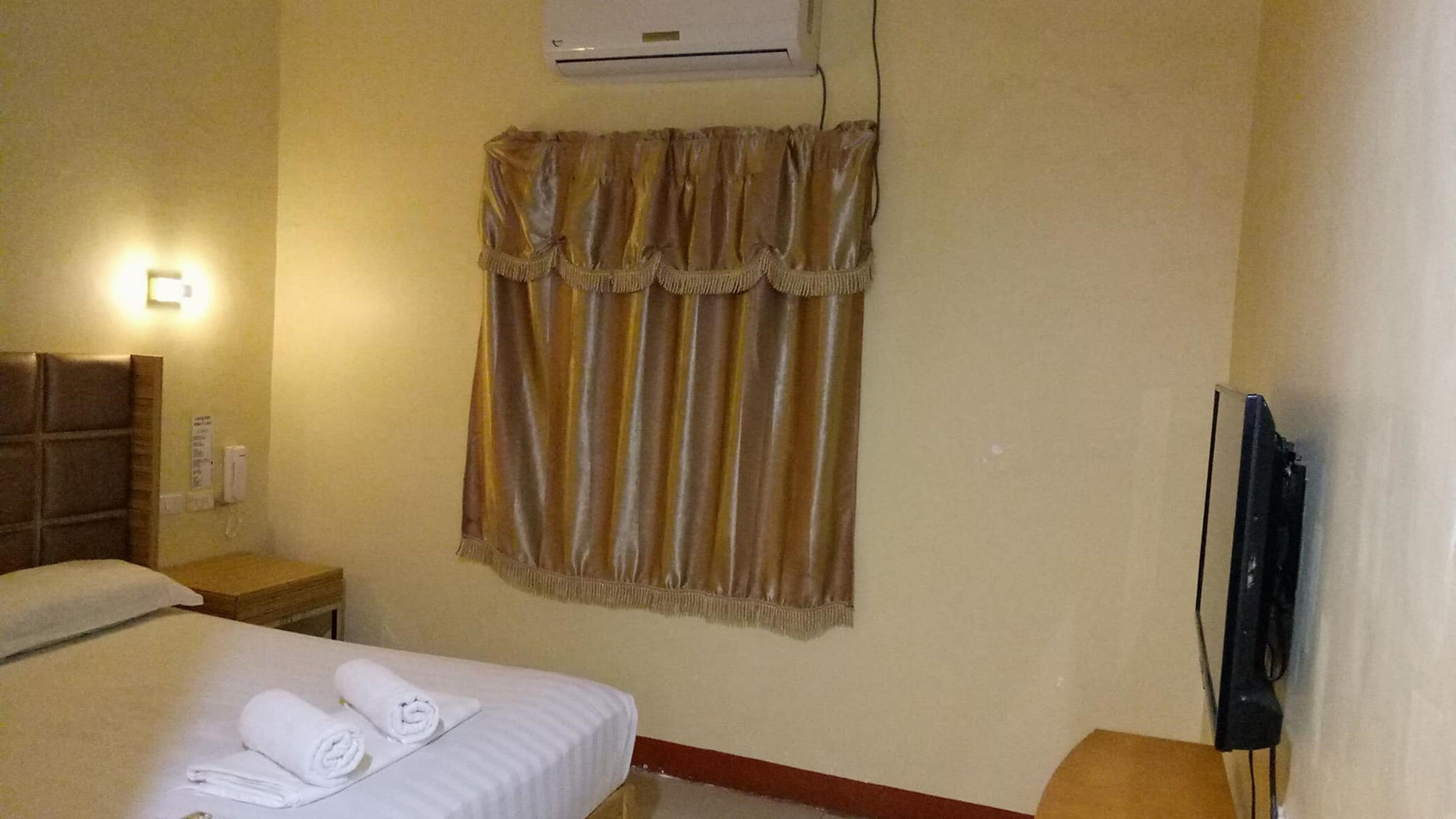 A hotel room with a bed, a bedside table, a wall-mounted air conditioner, and a television.