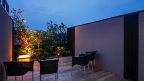 Un patio al aire libre con suelo de madera, dos sillas de mimbre negro y una lámpara de mesa encendida.