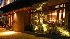 Un edificio con fachada de madera tradicional, una escalera que conduce a una entrada y un jardín bien iluminado con un pino.