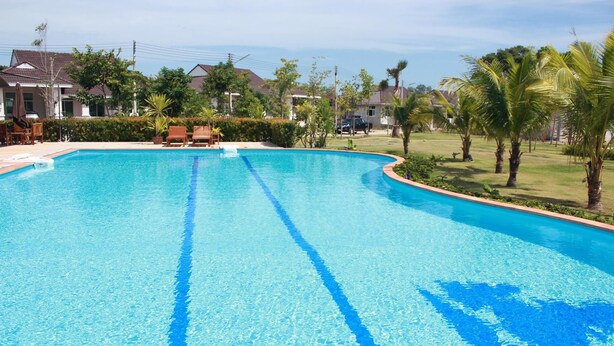 A clear swimming pool with a blue center line, surrounded by a grassy area with palm trees and houses in the background. - Suanson Paragon Green Villas (Rayong)