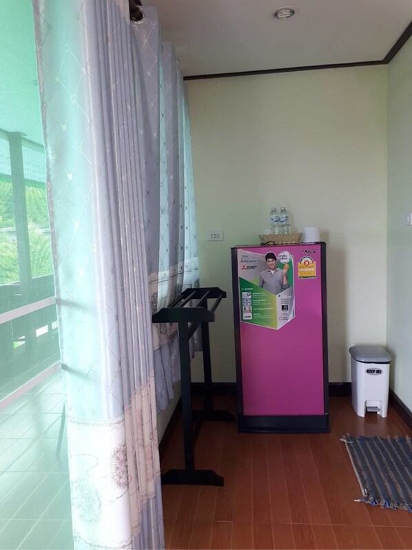 Um quarto com janela, porta, geladeira e uma pequena mesa, além de uma lixeira.
