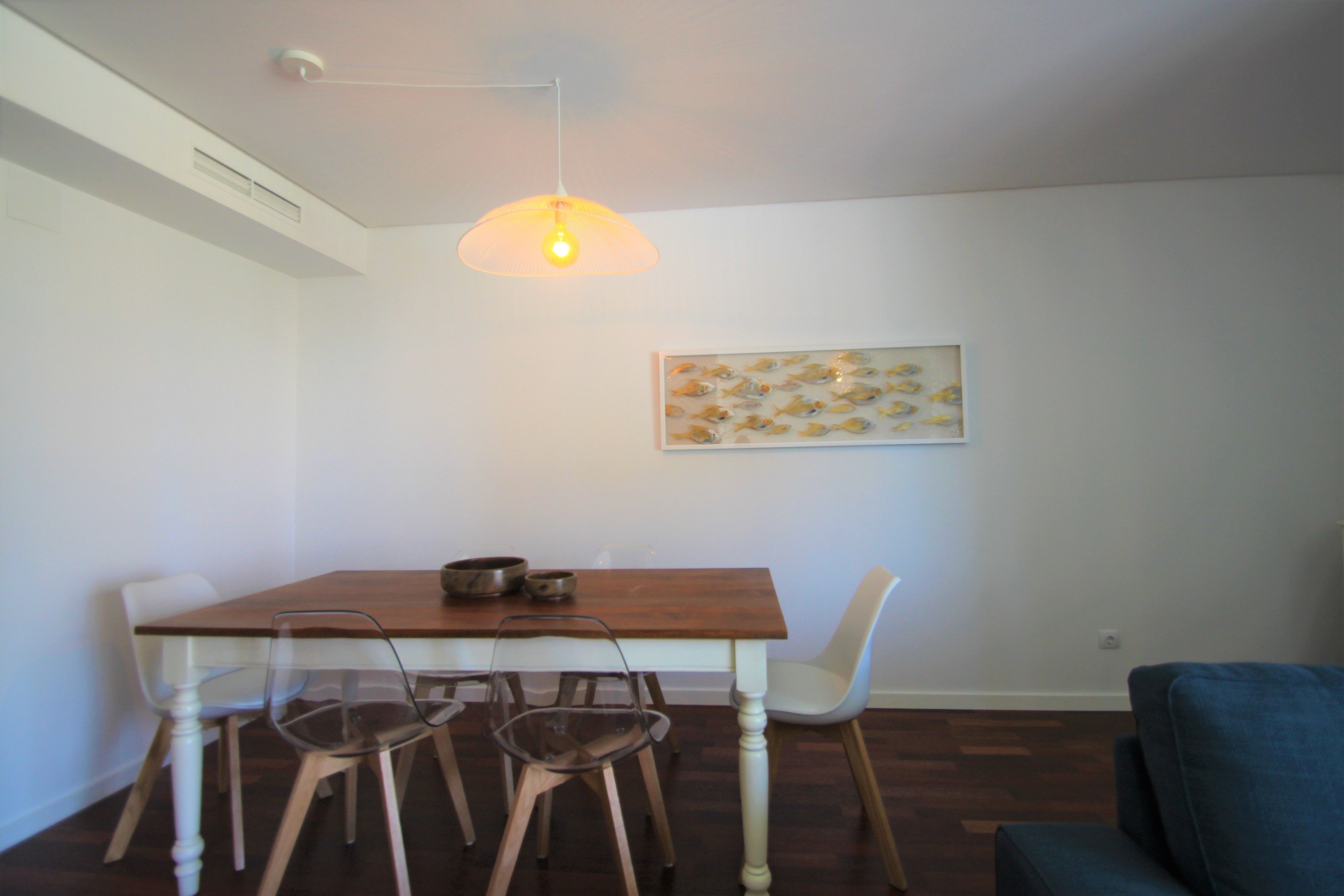 Un coin repas comprenant une table en bois, quatre chaises, un luminaire suspendu et un tableau représentant des poissons accroché au mur.