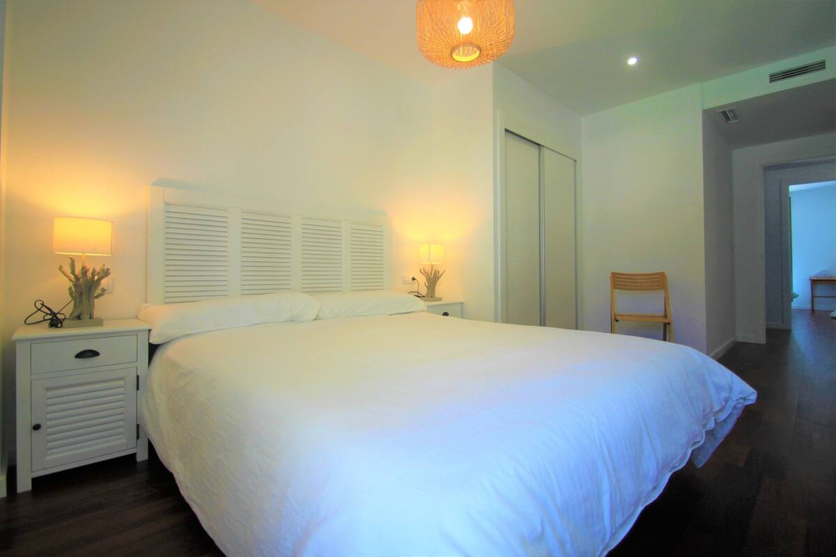A bedroom with a large bed, two bedside tables with lamps, a chair, and a door leading to another room.