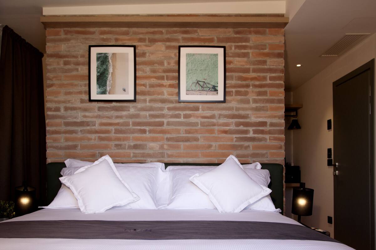 Habitación de hotel con pared de ladrillo, dos cuadros enmarcados, cama con ropa blanca y cabecera verde.