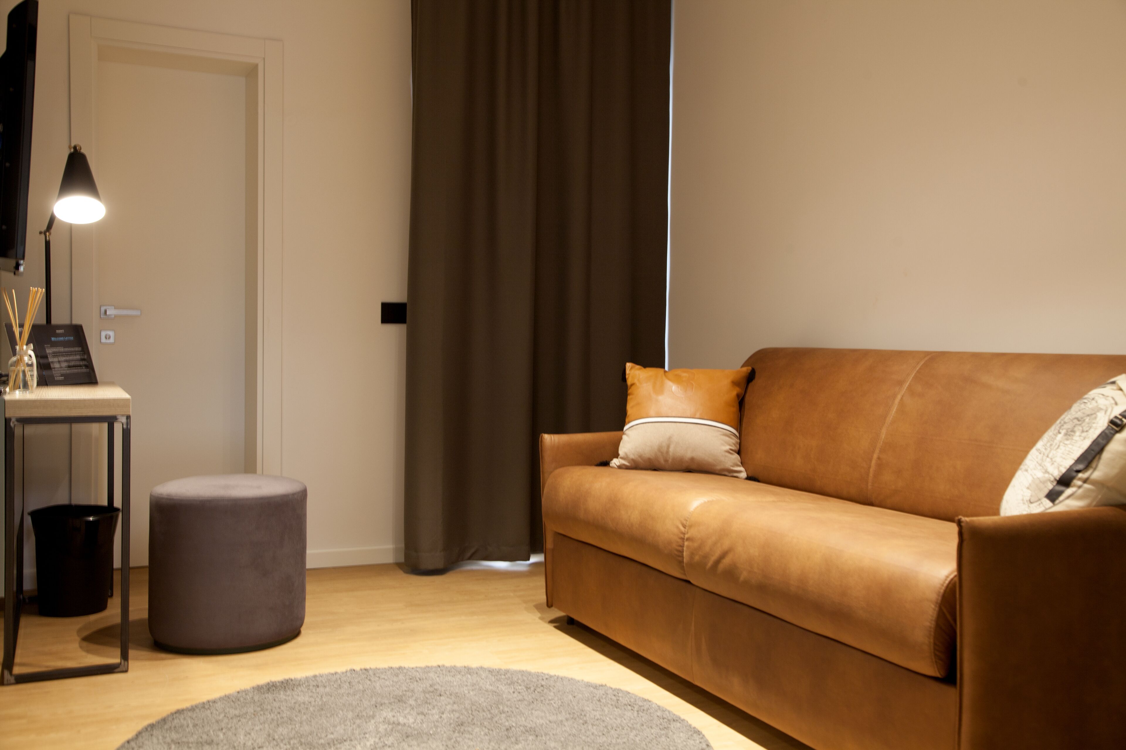 Una sala moderna con un sofá de cuero marrón, un reposapiés gris, una mesita con una lámpara y una alfombra gris.