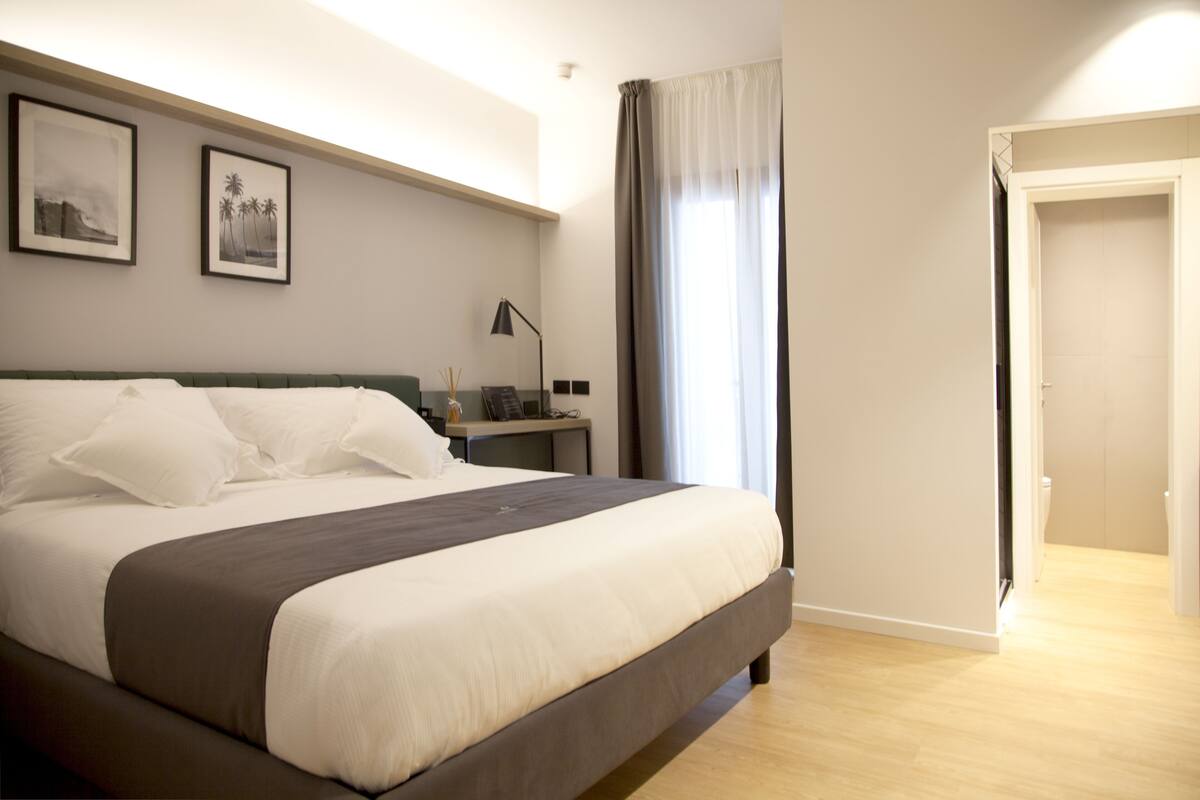 Una cama bien hecha con ropa de cama blanca y oscura, una mesita de noche con una lámpara y dos cuadros en la pared.