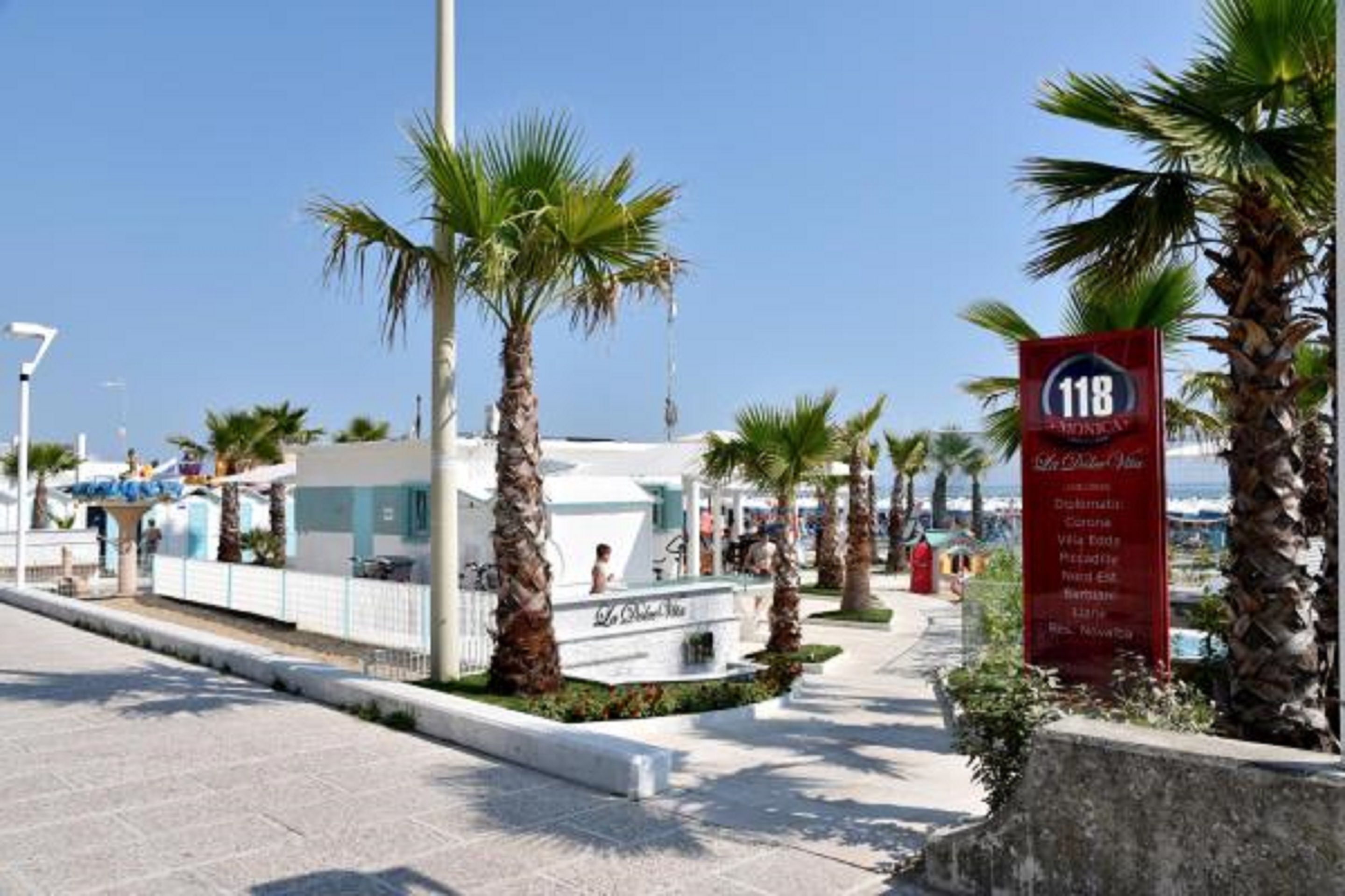 A coastal walkway with palm trees, a sign for '118', and a building with a white facade.