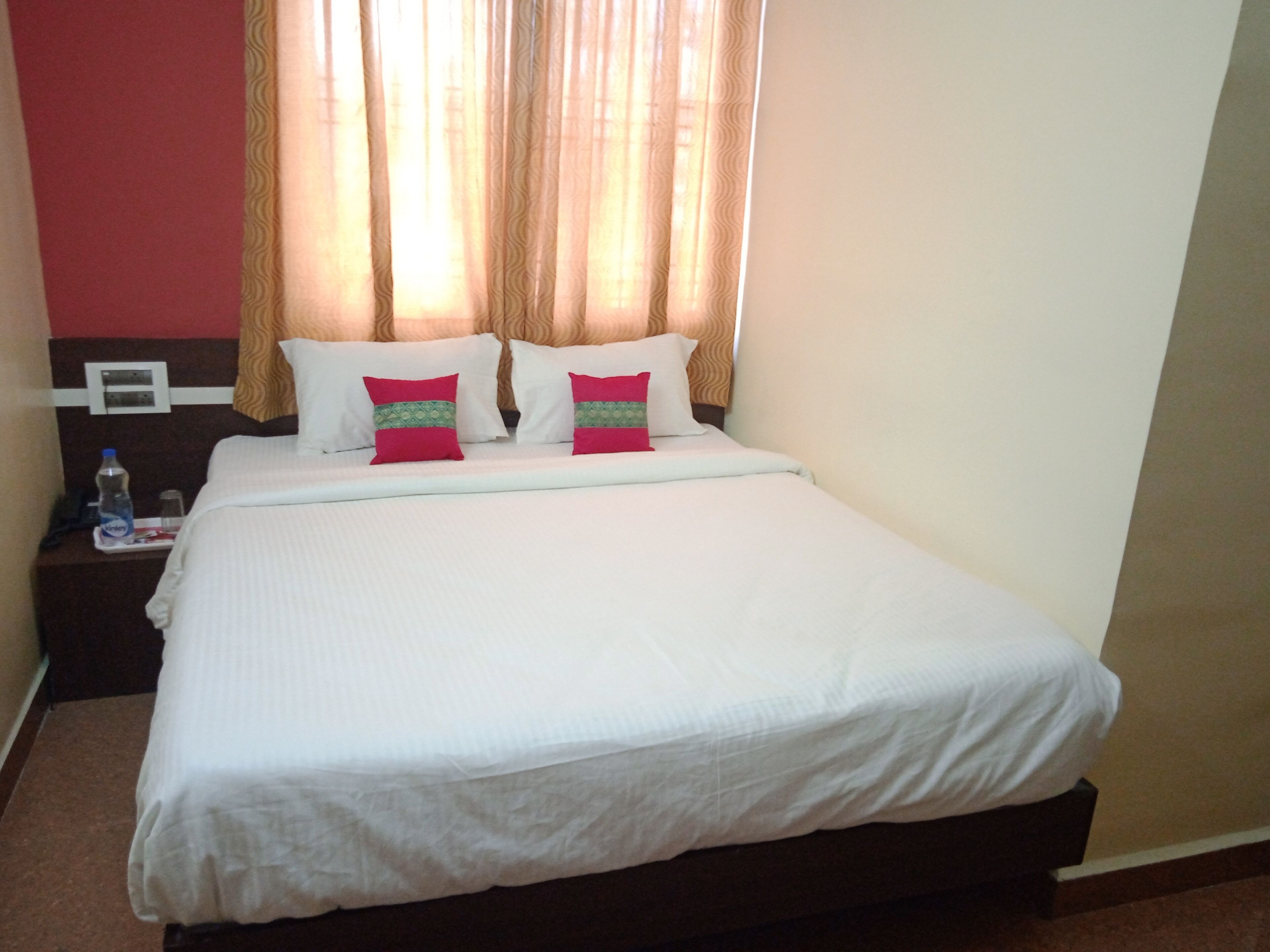 Cama bem arrumada com roupa de cama branca e duas almofadas decorativas, mesa de cabeceira com garrafa de água e telefone, e janela com cortinas bege.