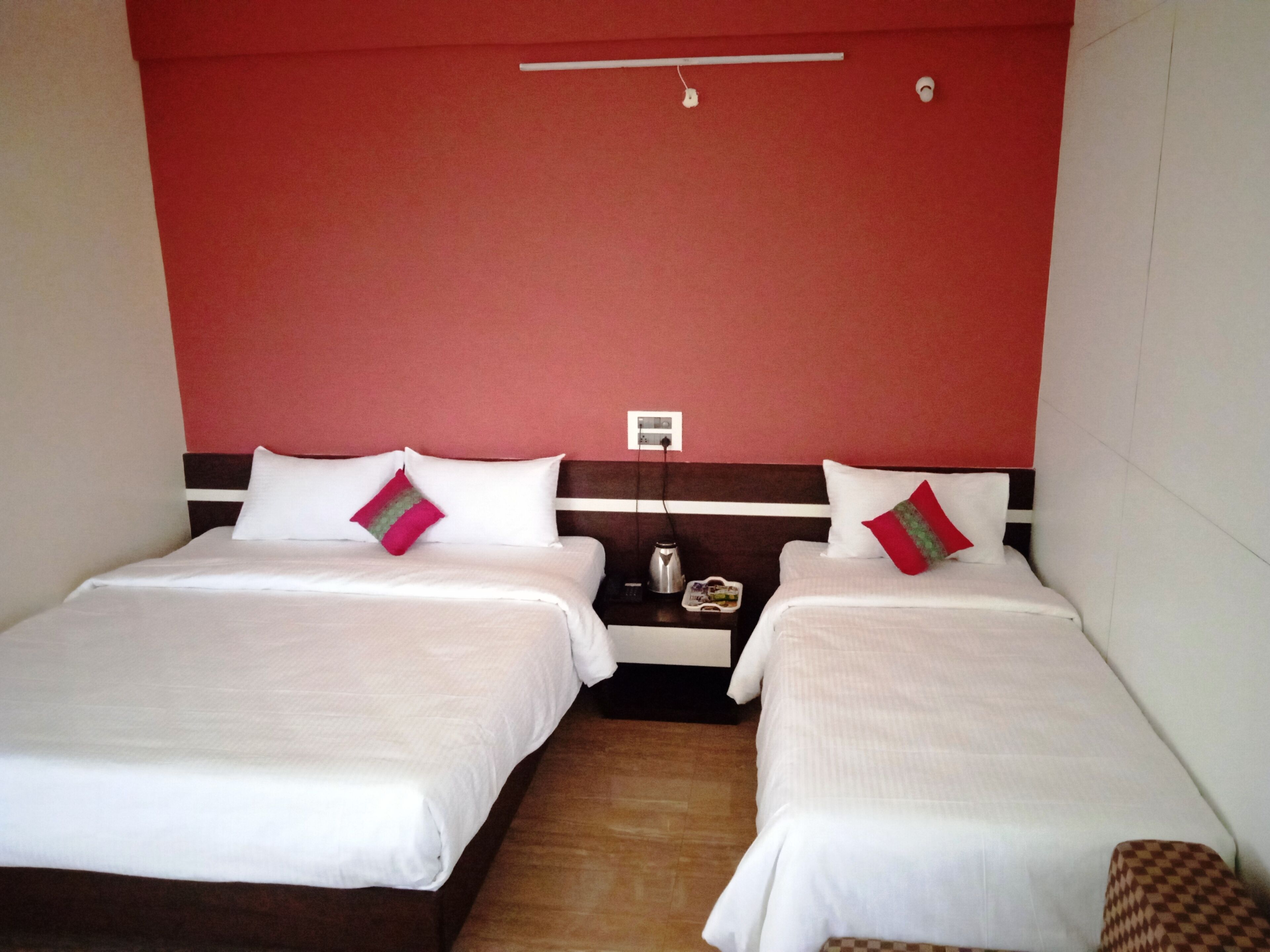 Habitación de hotel con dos camas, cada una con ropa blanca y una almohada roja. Hay una mesita de noche con una tetera y una pequeña mesa entre las camas. Las paredes están pintadas de rojo y hay un accesorio de luz blanca en el techo sobre las camas.