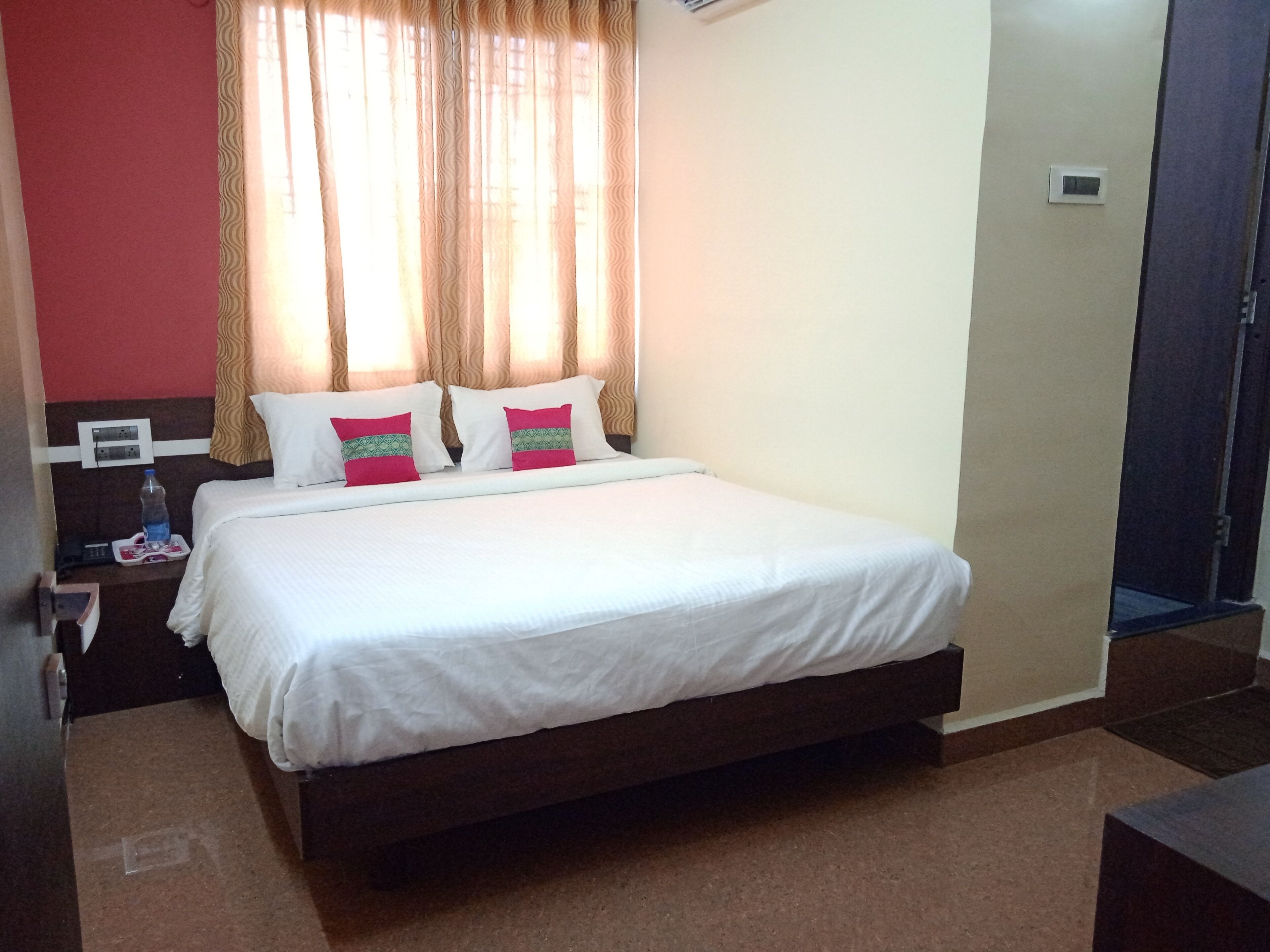 A hotel room with a large bed, two pillows, a water bottle on the nightstand, and a thermostat on the wall.