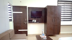 A hotel room with a bed, a sofa, a small table, a TV mounted on a cabinet, and a window with blinds. - Sagar Resort (Chhatrapati Sambhajinagar)