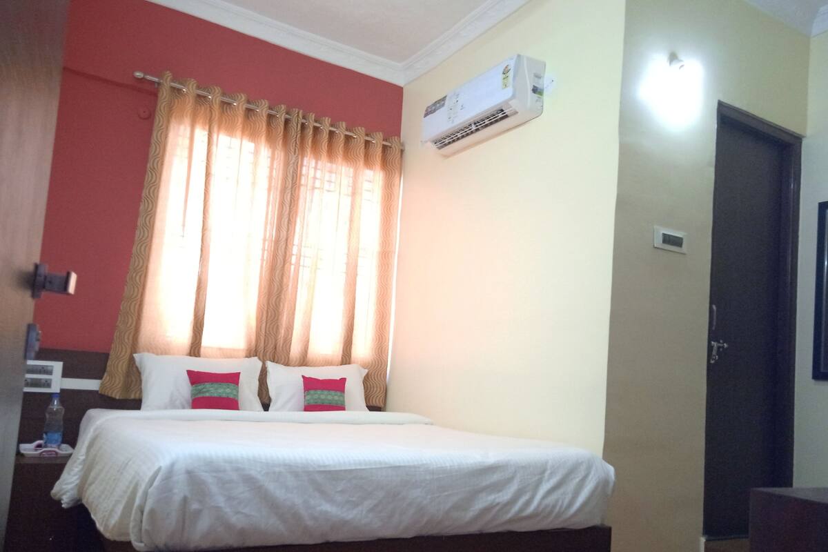 A hotel room with a bed, curtains, an air conditioning unit, and a door.