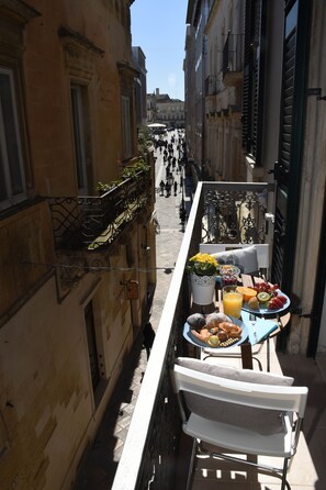 Una estrecha calle urbana con una mesa en un balcón dispuesta con alimentos para el desayuno, con vista a una calle concurrida.