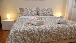Un lit bien fait, recouvert d’une couvre-lit à motifs, deux serviettes pliées, maintenues par un ruban, et des lampes de chevet posées sur des tables de nuit blanches.