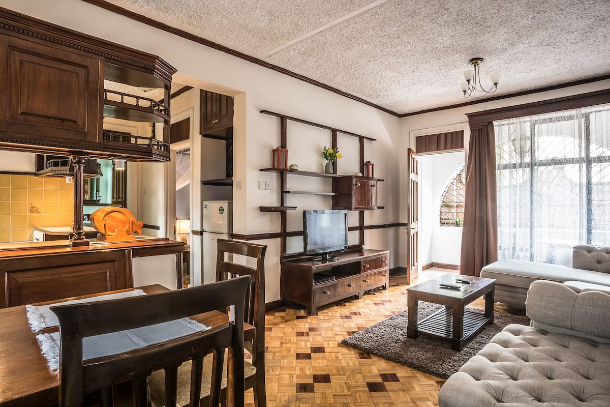 A living room with a wooden dining table, chairs, a sofa, a television on a wooden stand, and a window with curtains.