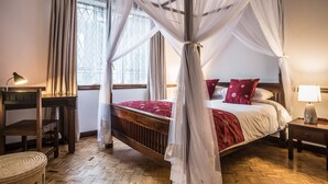 A bedroom with a four-poster canopy bed, a wooden headboard, a bedside table with a lamp, and a window with sheer curtains. - The Grand Gables (Nairobi)