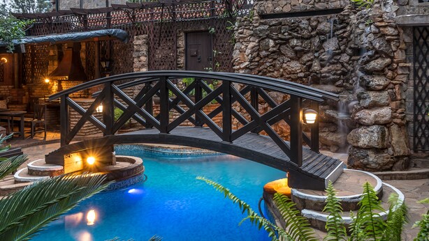 A pool area with a stone wall, a wooden bridge, and a seating area with tables and chairs. - The Grand Gables (Nairobi)