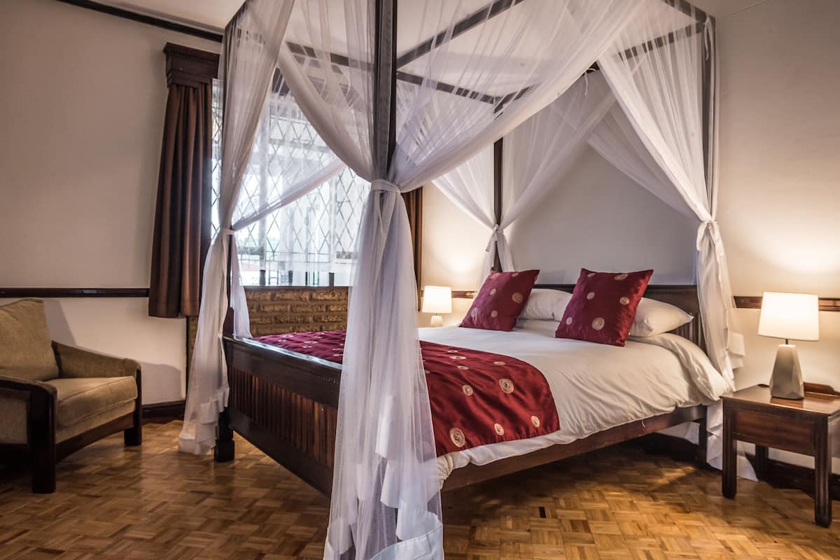 A four-poster canopy bed with a wooden headboard, two bedside tables with lamps, a chair, and a view of a building through a window.