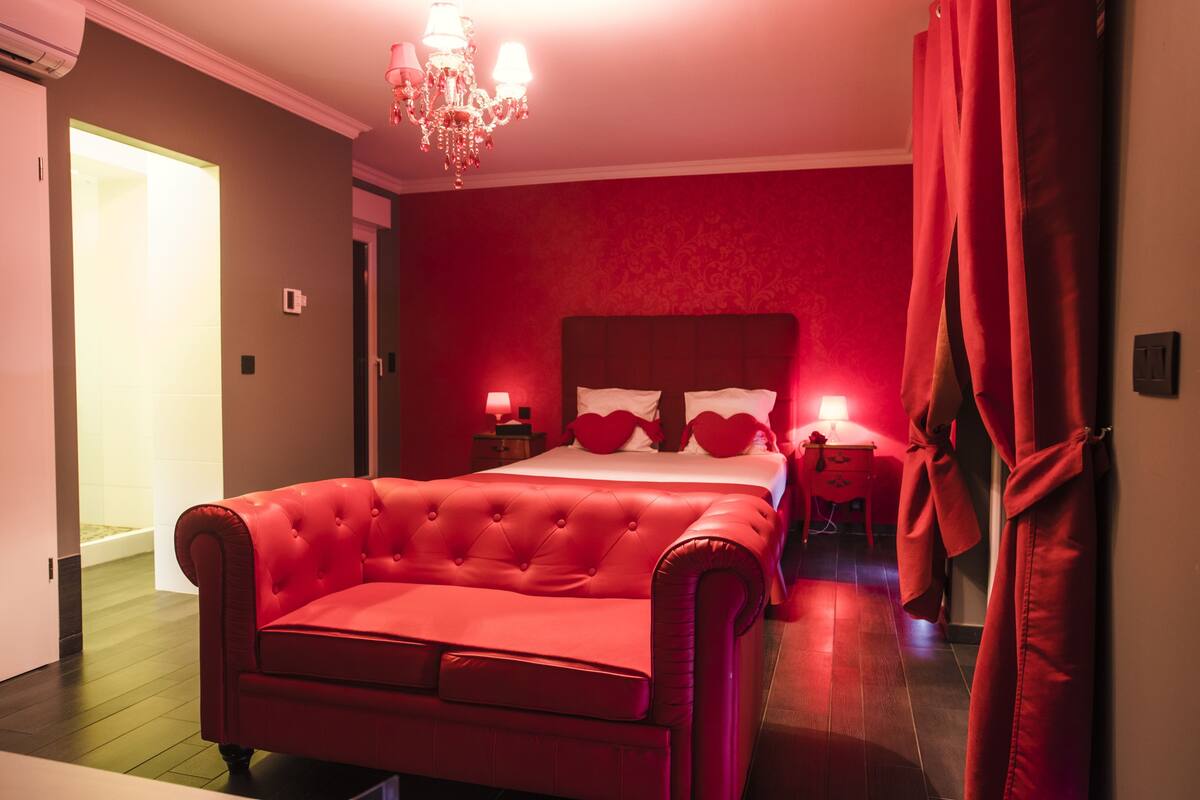 Quarto com decoração em tons de vermelho, cama com cabeceira estofada, luminárias de apoio e um lustre.