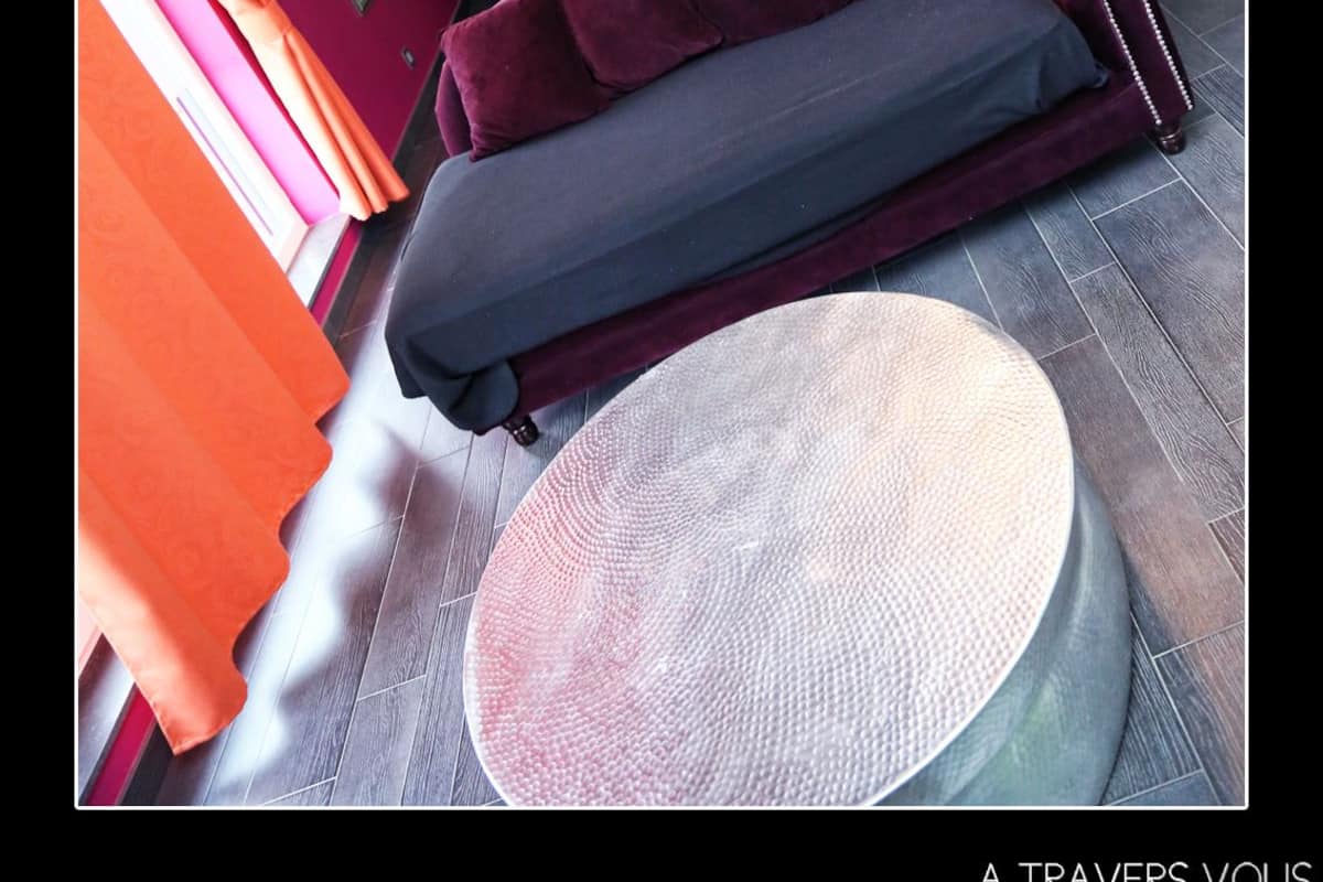Quarto com cabeceira roxa, cama com edredom cinza e roxo, mesa lateral redonda e lustre.
